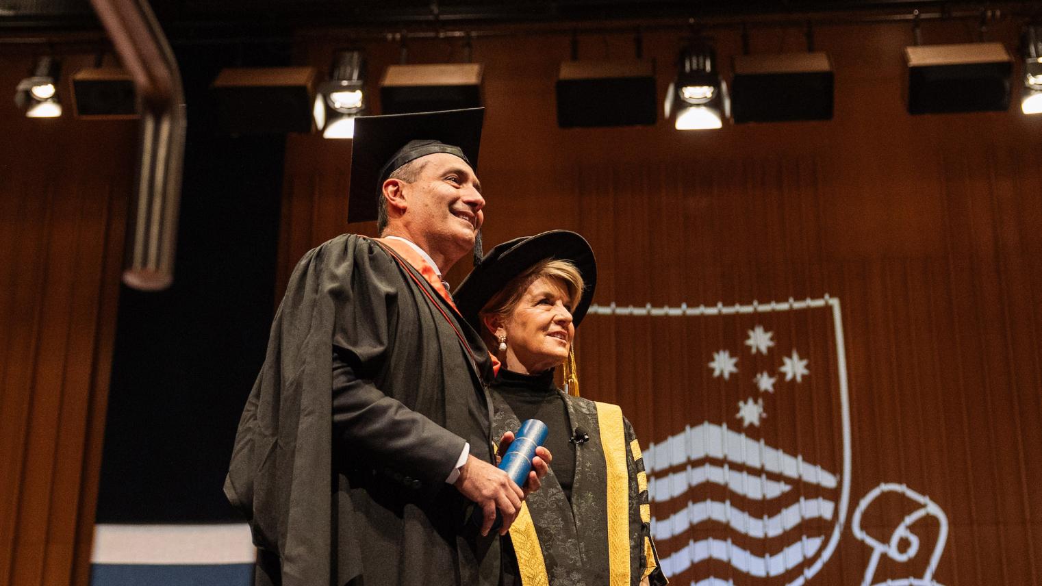 Steven Kourras and Chancellor of the Australian National University Julie Bishop at the 2023 Mid-Year Graduation Steven Kourras standing on stage with Julie Bishop in graduation robes, holding his Testamur